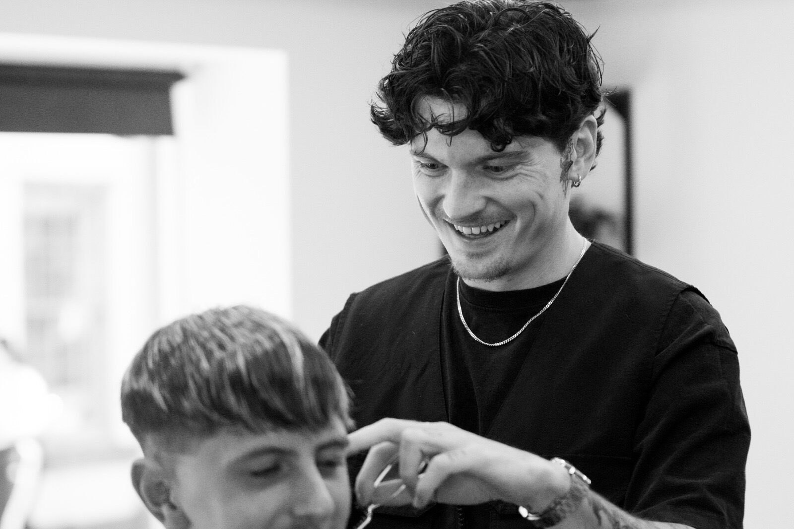 Black and white photo of a barber smiling as he works on a client's fringe